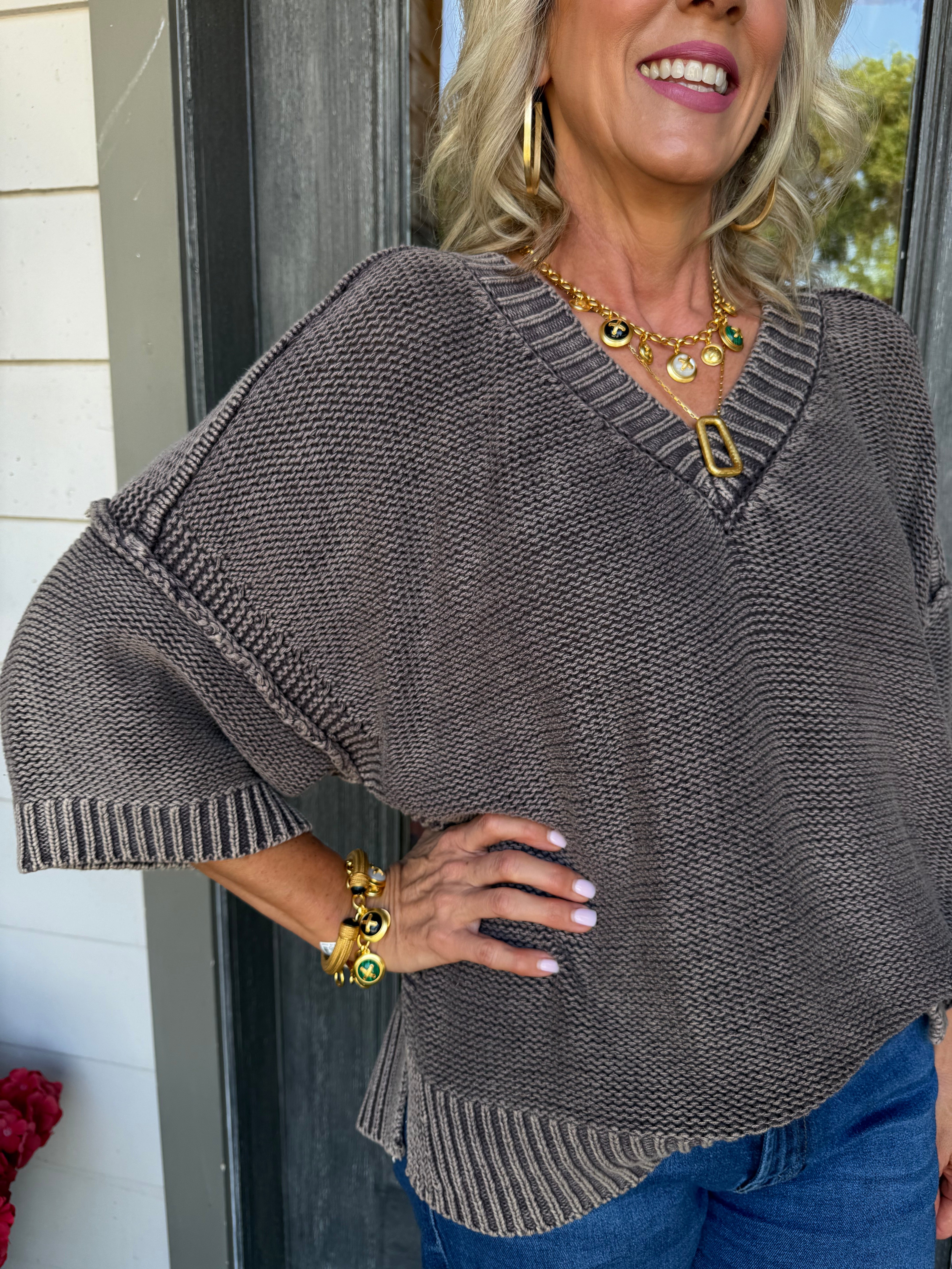 A woman wearing a brown textured knit sweater with a V-neck and wide three-quarter sleeves, paired with blue jeans and gold jewelry, standing in front of a doorway.