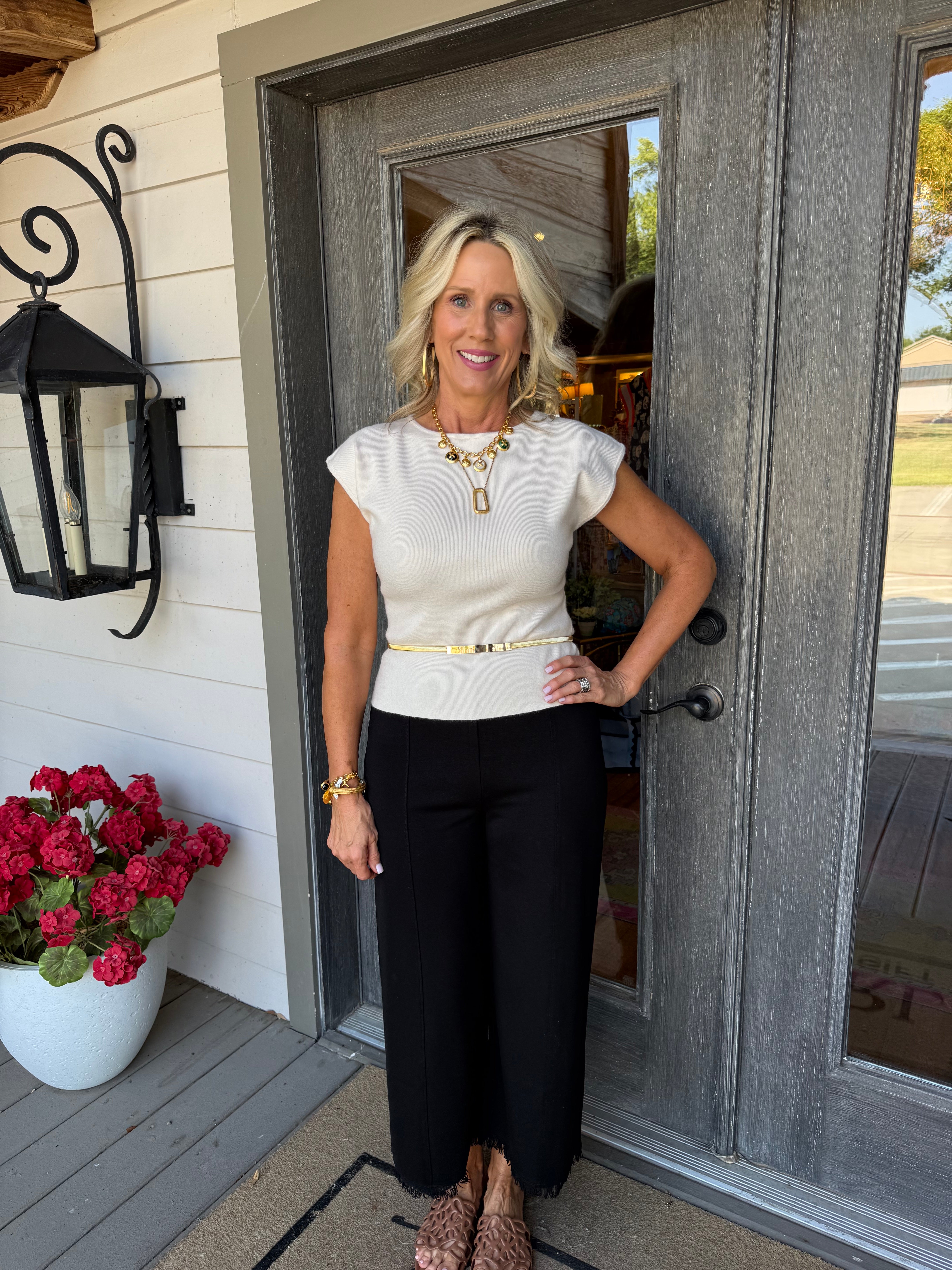 A woman stands on a front porch wearing a cream short-sleeve top with a thin gold belt, black wide-leg pants, and leopard-print heels. She is positioned in front of a gray double door with a black wall lantern and a potted plant of red flowers to her left.
