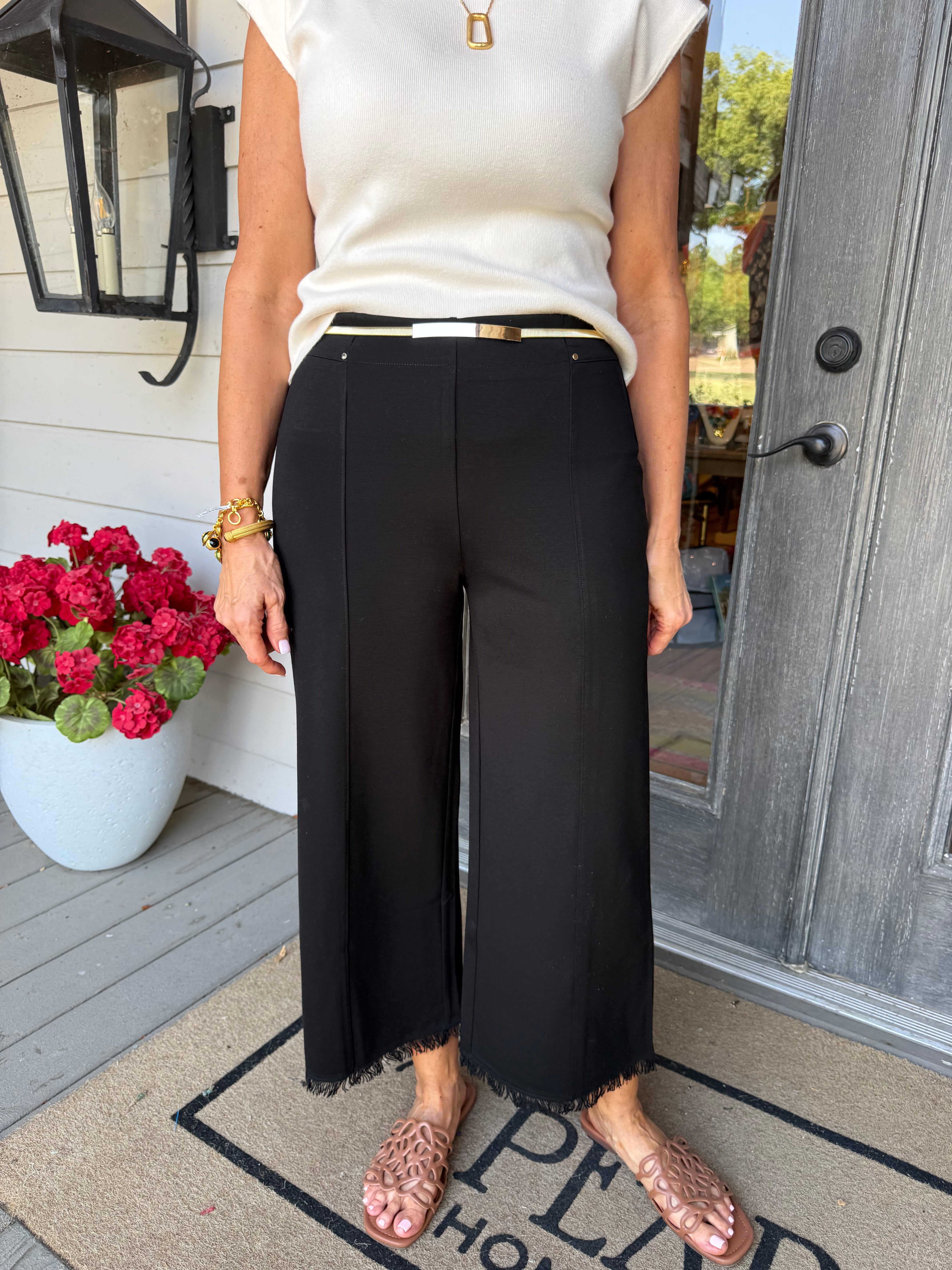 A person wearing black wide-leg cropped pants with a thin gold belt, paired with a cream sleeveless top and tan flat sandals, standing on a front porch.
