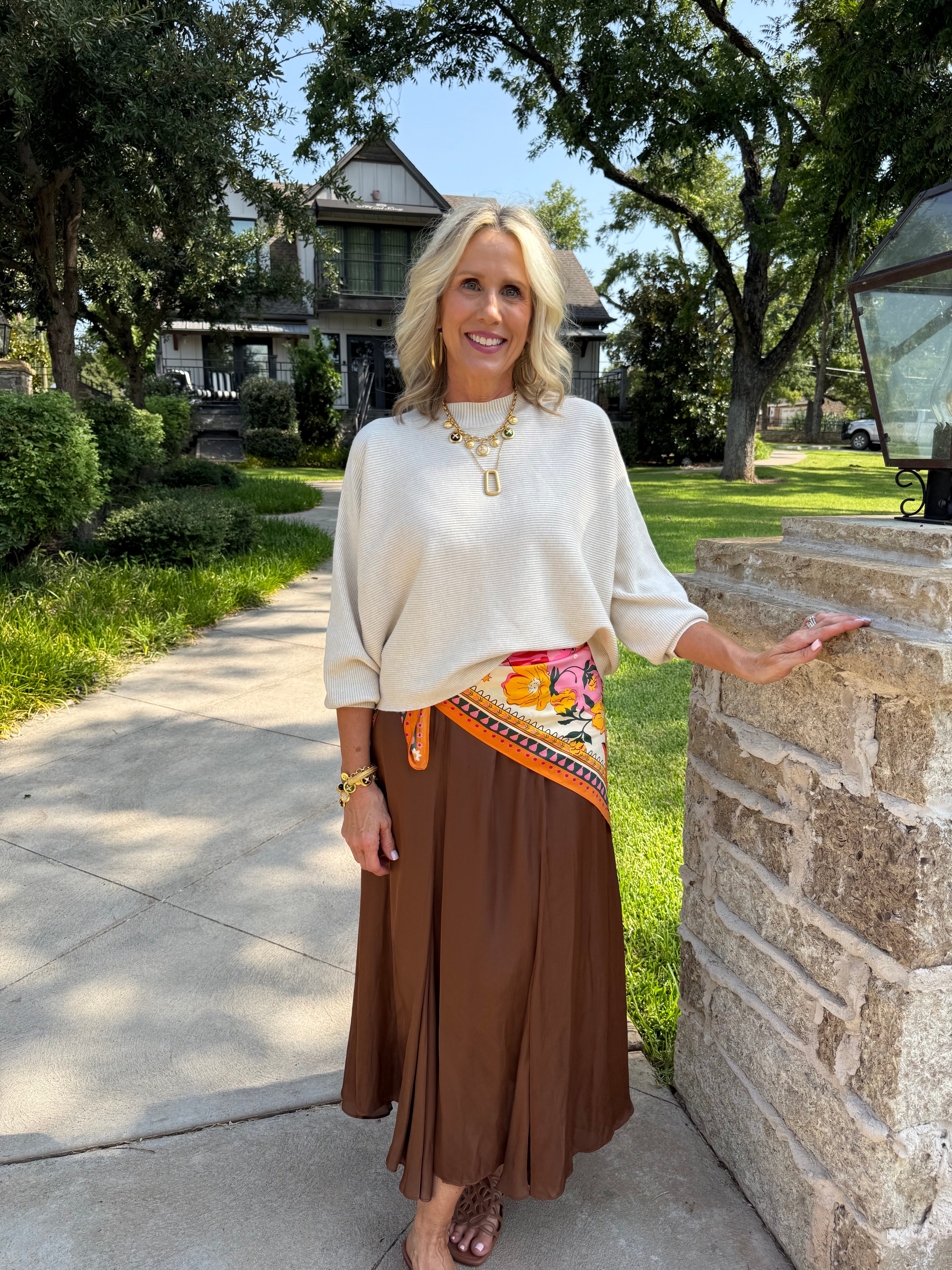 A woman stands outdoors on a stone walkway beside a stone pillar, wearing a cream-colored long-sleeve top, a brown pleated maxi skirt, and a colorful patterned wrap belt. She is accessorized with gold jewelry and sandals, with a suburban house and trees in the background.