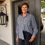 A smiling woman with short brown hair stands on a porch wearing a navy and white gingham button-down shirt and dark navy pants.