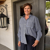 A smiling woman with short brown hair stands on a porch wearing a navy and white gingham button-down shirt and dark navy pants.