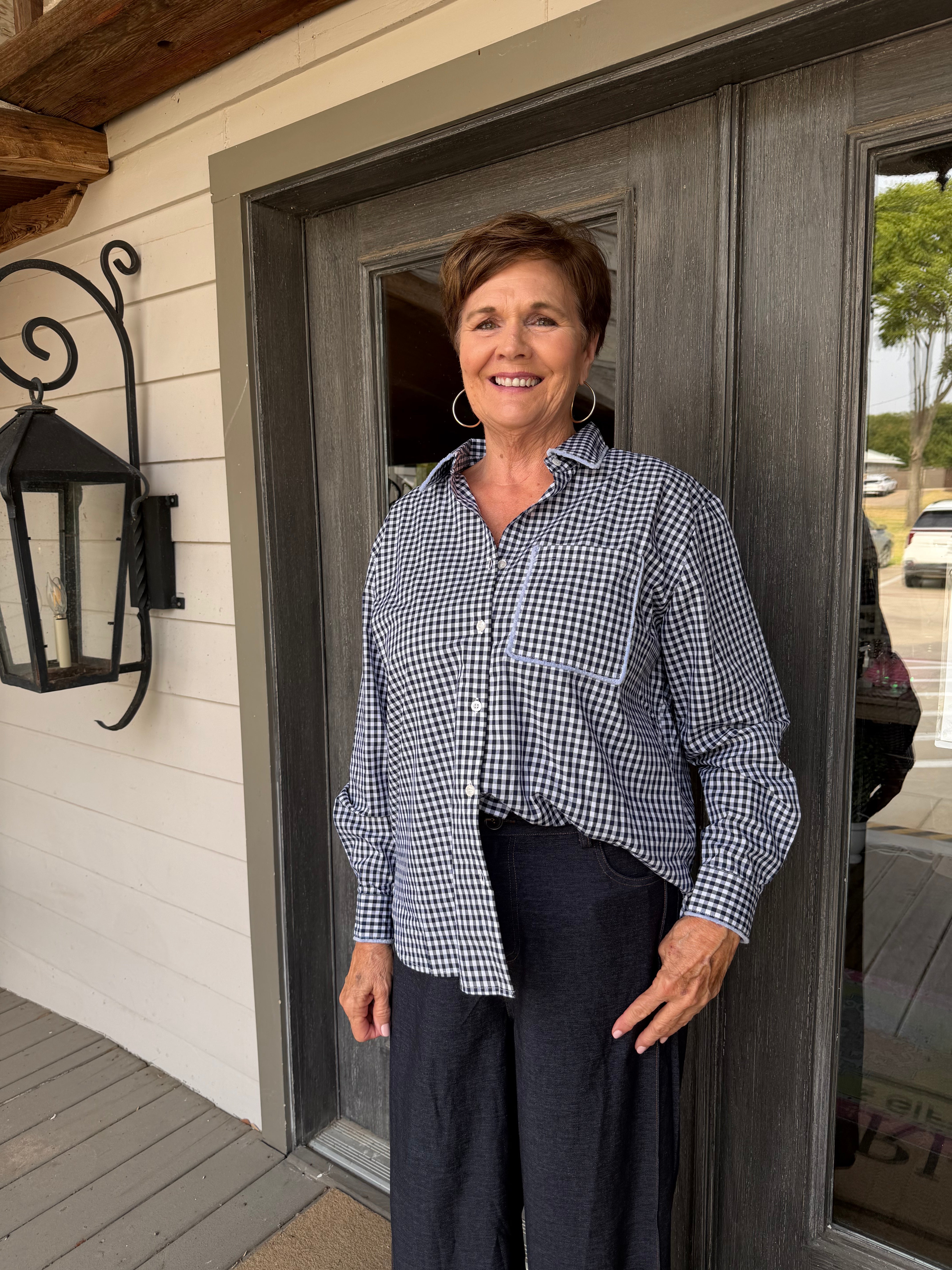 A smiling woman with short brown hair stands on a porch wearing a navy and white gingham button-down shirt and dark navy pants.