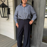 A woman stands on a front porch wearing a blue and white gingham button-down shirt tucked into dark navy wide-leg trousers, paired with white sneakers.