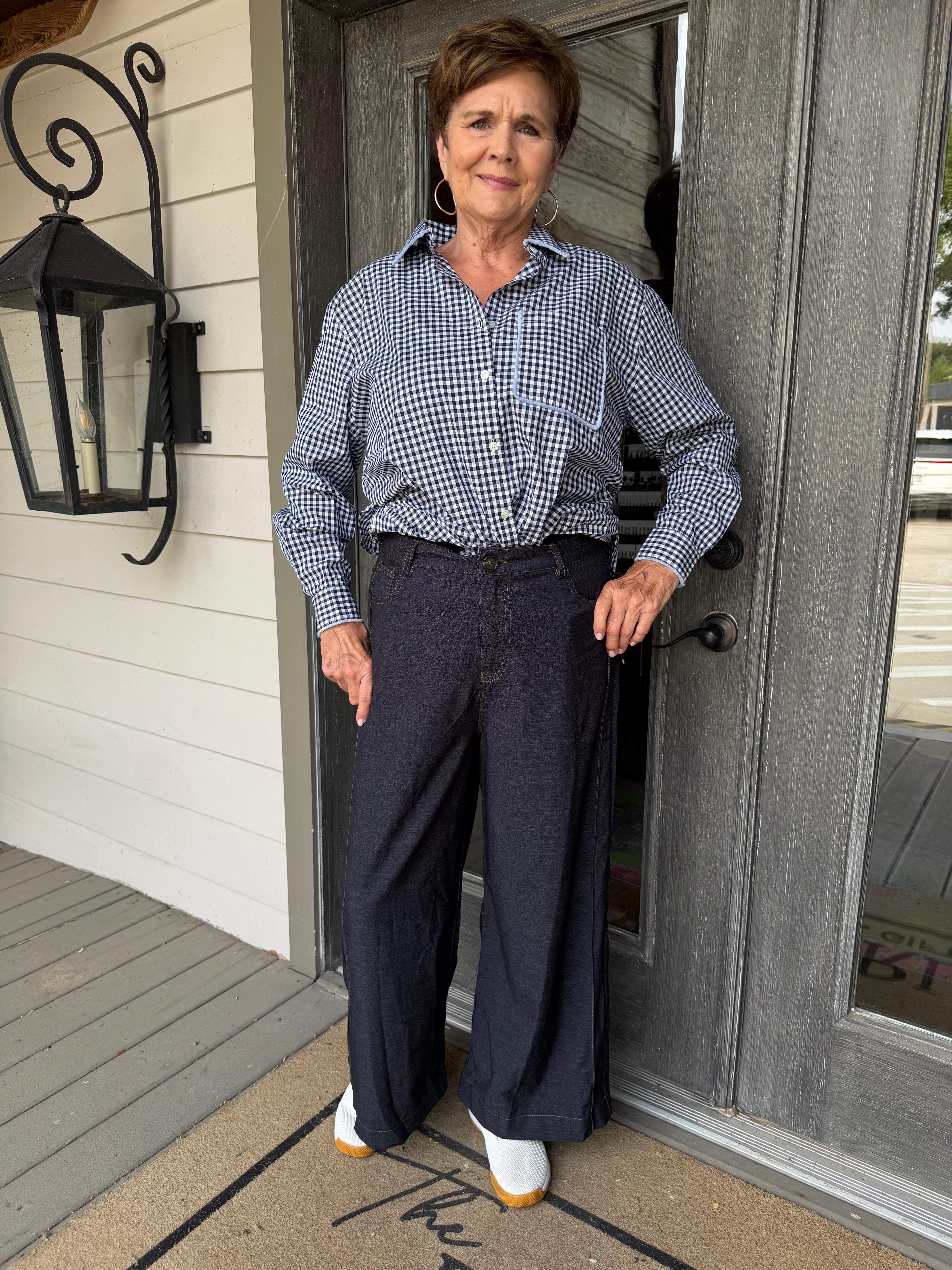 A woman stands on a front porch wearing a blue and white gingham button-down shirt tucked into dark navy wide-leg trousers, paired with white sneakers.