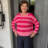 A woman stands on a porch wearing a pink and magenta striped knit sweater and dark navy pants, posing in front of a gray wooden door.