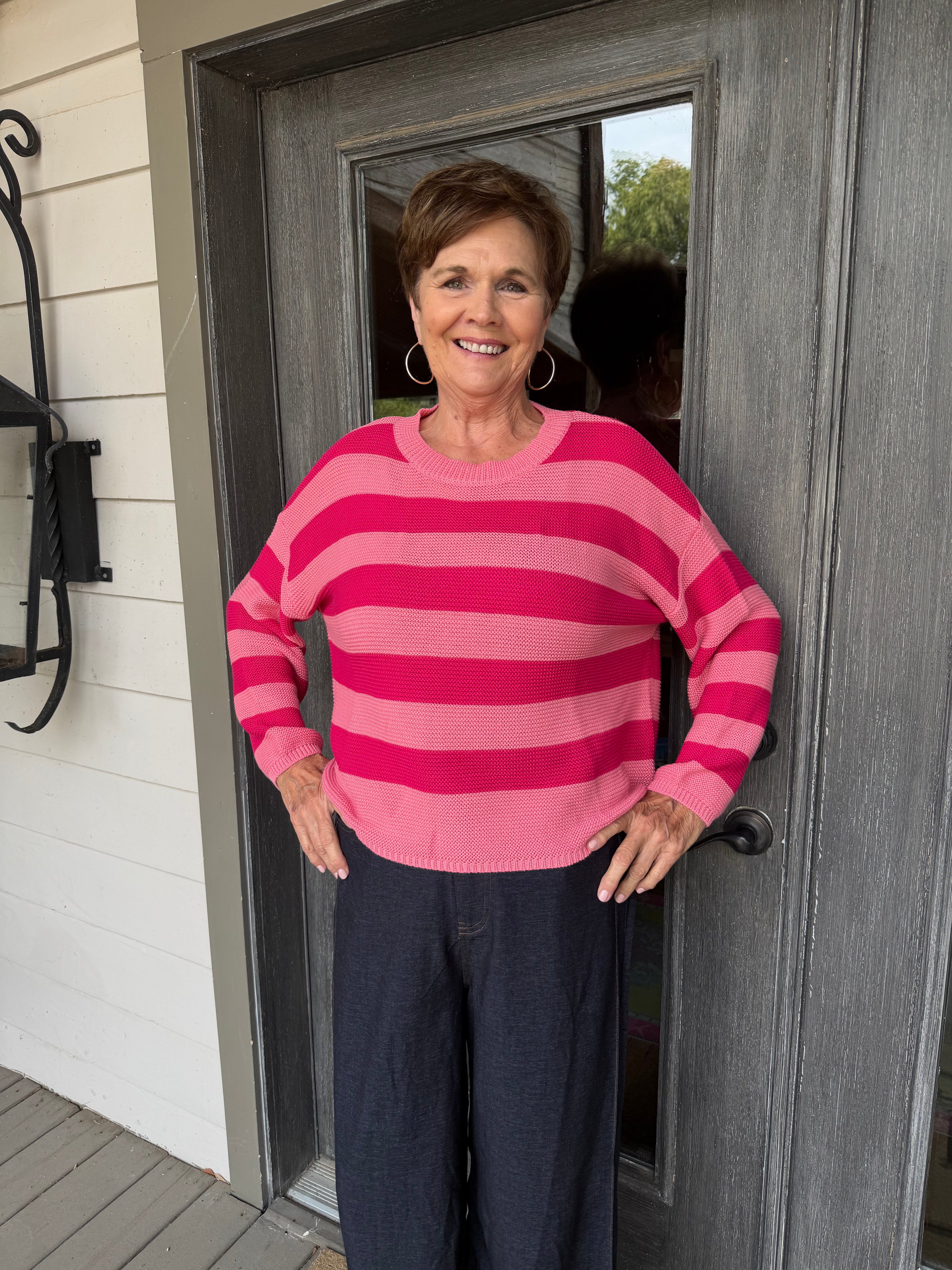 A woman stands on a porch wearing a pink and magenta striped knit sweater and dark navy pants, posing in front of a gray wooden door.