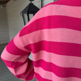 A close-up of a person wearing a pink and magenta striped knit sweater, photographed from behind on a porch with a lantern and siding in the background.