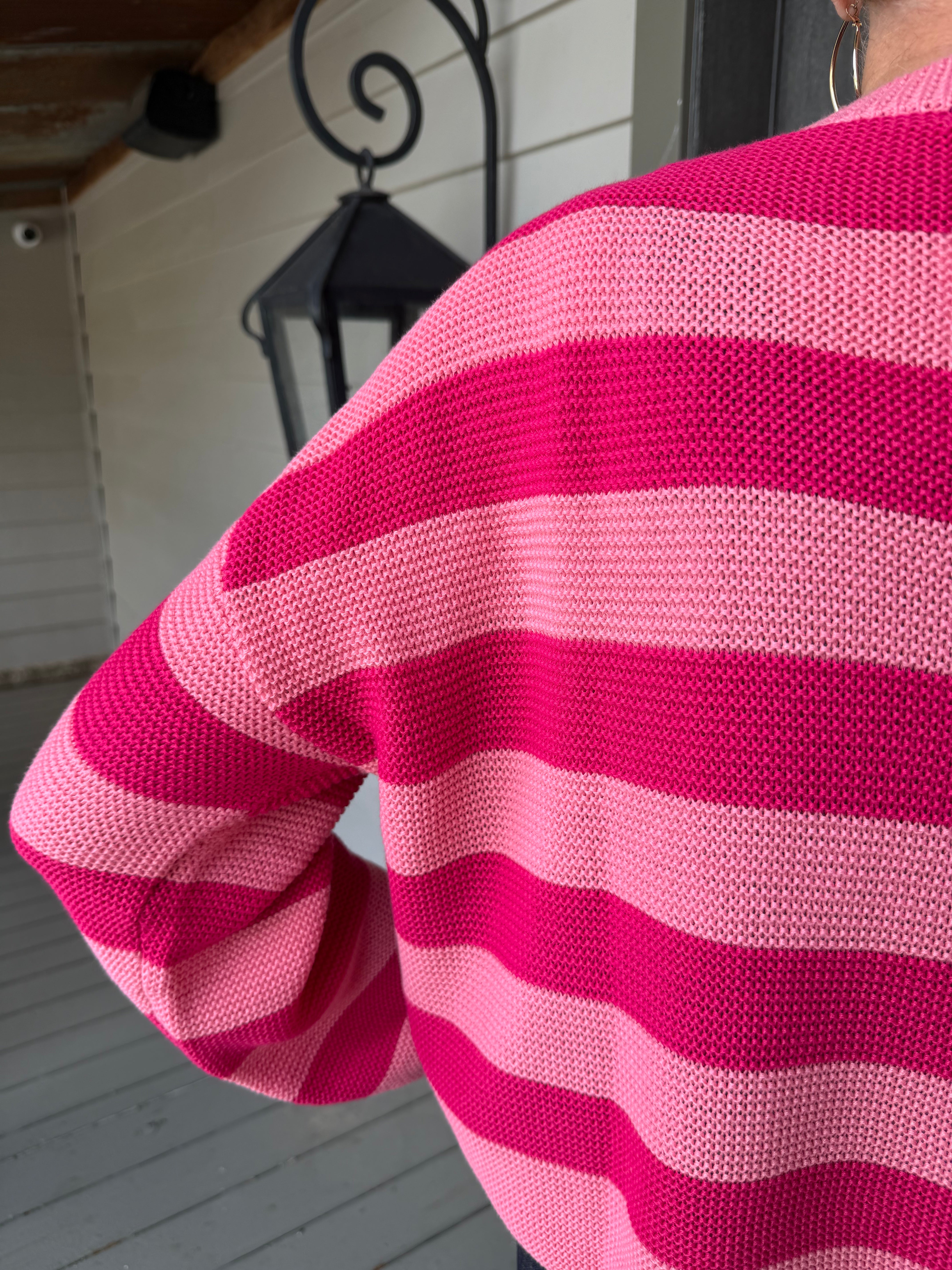 A close-up of a person wearing a pink and magenta striped knit sweater, photographed from behind on a porch with a lantern and siding in the background.