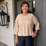 A woman stands on a porch wearing a beige cable-knit three-quarter sleeve top and dark navy pants.