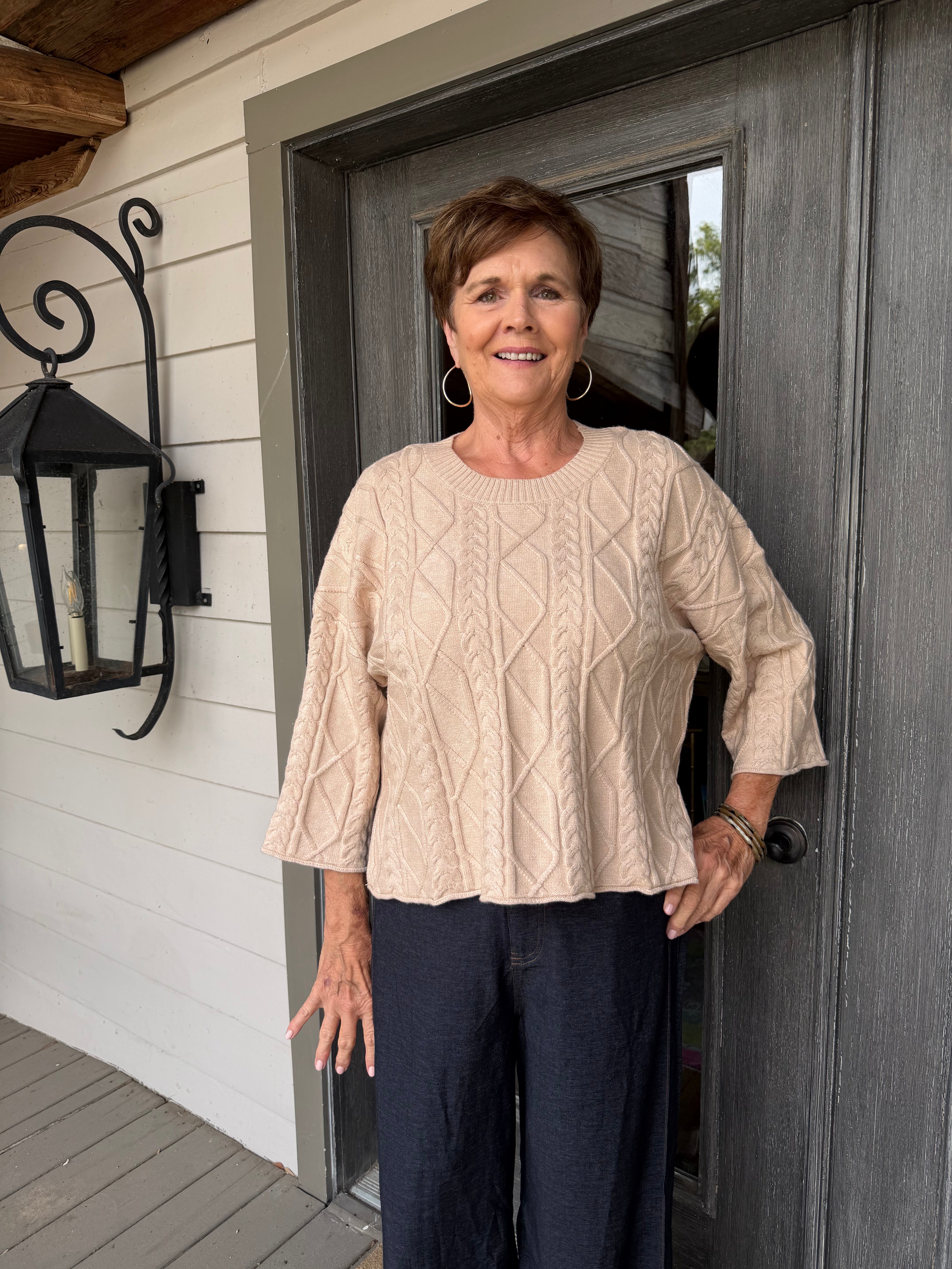 A woman stands on a porch wearing a beige cable-knit three-quarter sleeve top and dark navy pants.
