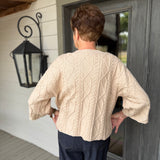 A person viewed from behind wearing a cream cable-knit cropped sweater and dark navy pants, standing on a porch in front of a gray door.