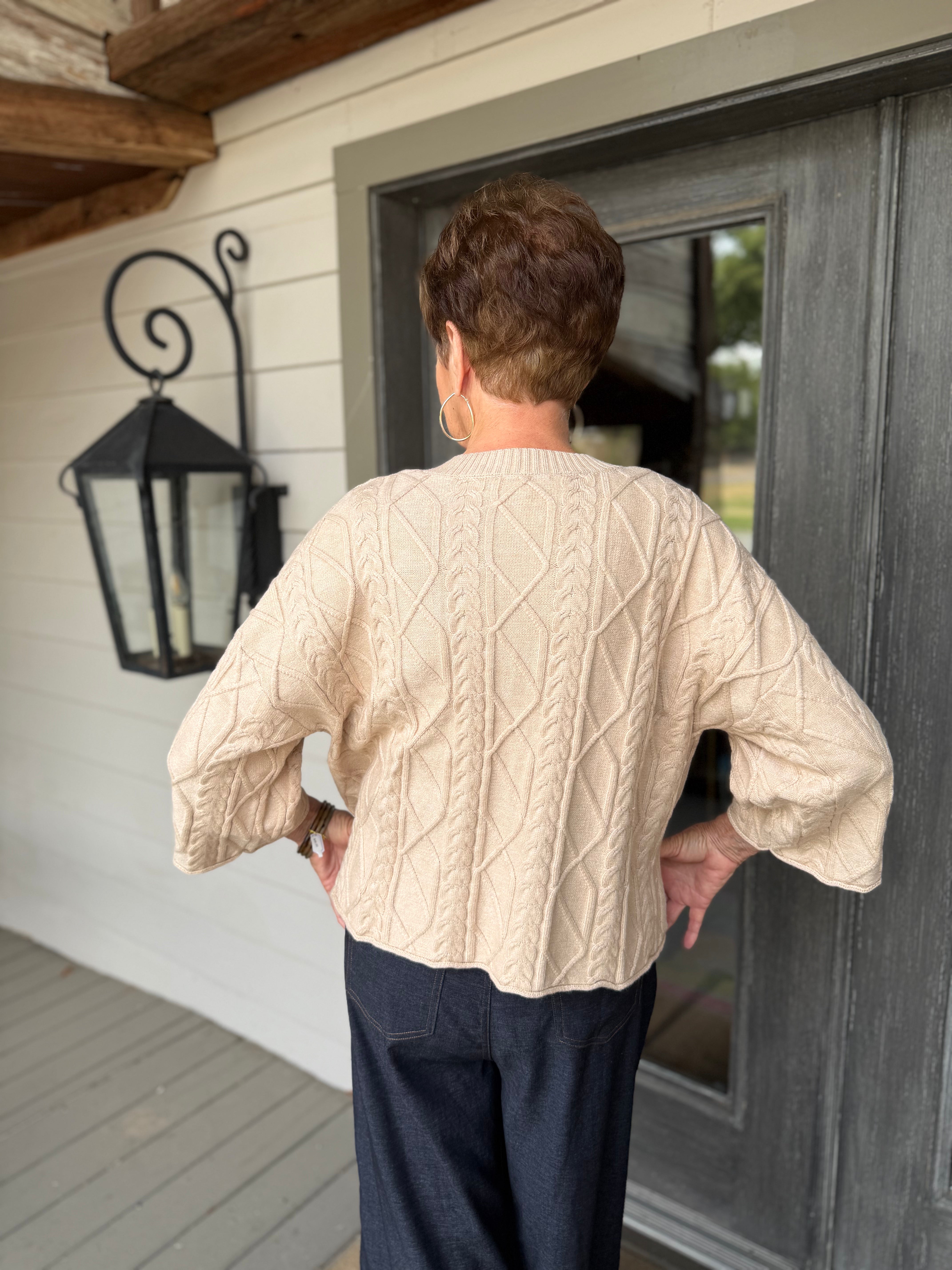 A person viewed from behind wearing a cream cable-knit cropped sweater and dark navy pants, standing on a porch in front of a gray door.