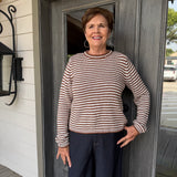 A smiling older woman stands on a porch in front of a gray double door, wearing a brown and cream horizontally striped long-sleeve knit sweater and dark navy pants.