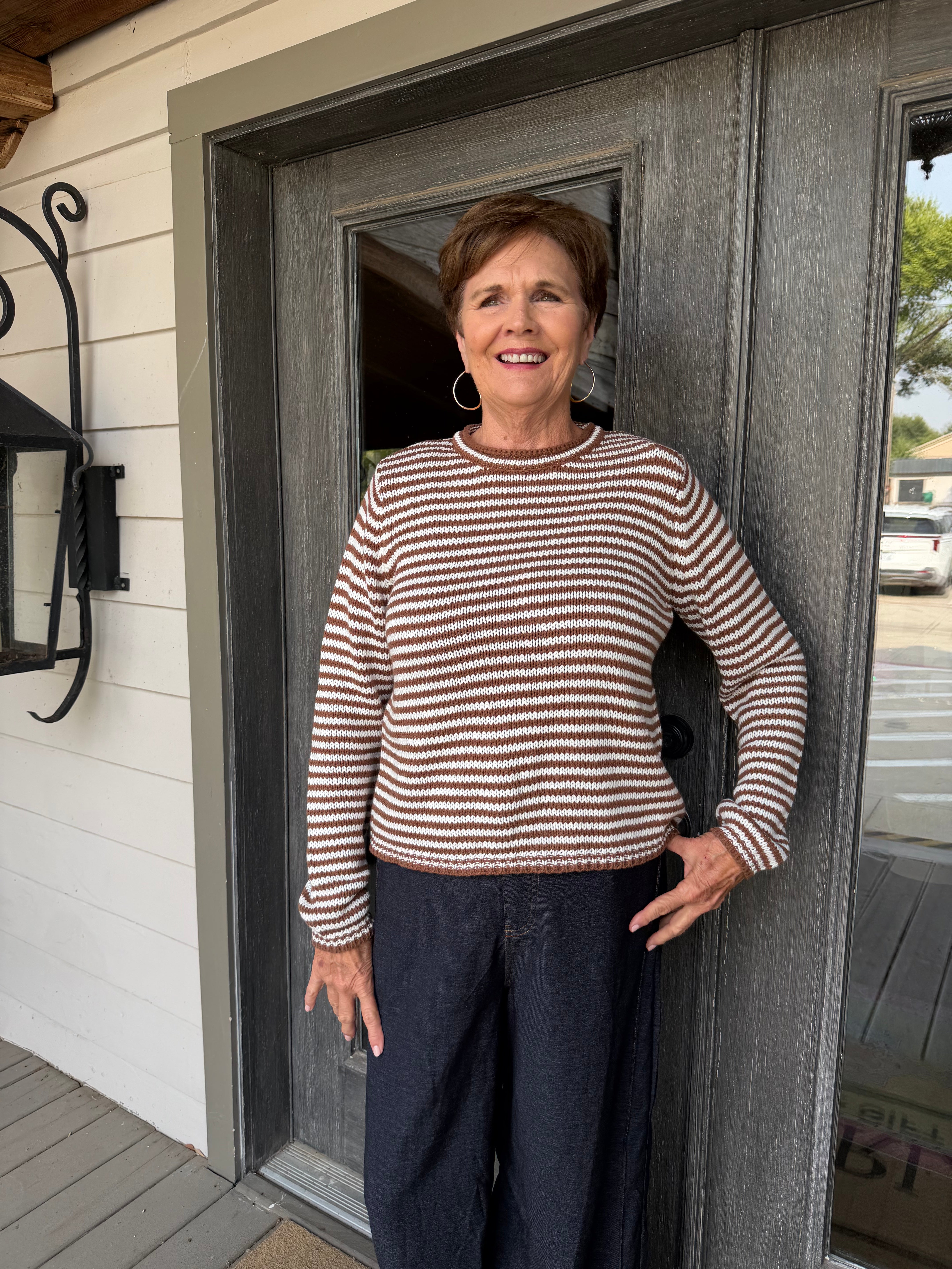 A smiling older woman stands on a porch in front of a gray double door, wearing a brown and cream horizontally striped long-sleeve knit sweater and dark navy pants.
