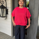 A woman wearing a bright pink short-sleeve knit sweater and dark wide-leg pants stands on a front porch in front of a gray door.