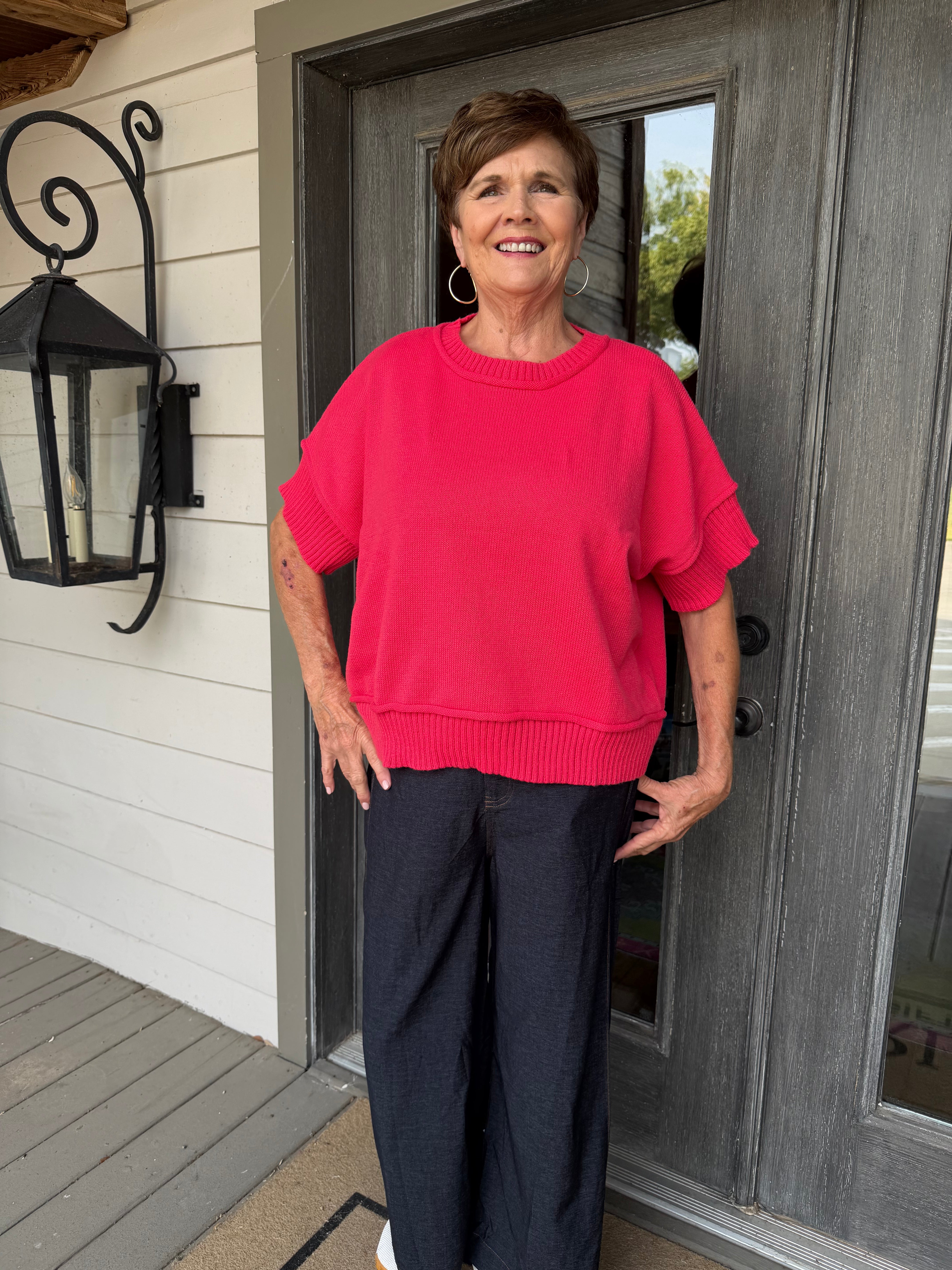 A woman wearing a bright pink short-sleeve knit sweater and dark wide-leg pants stands on a front porch in front of a gray door.