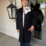 A woman stands on a porch wearing a long black cardigan over a white blouse with a cream bow, navy wide-leg pants, and flat sandals. The background shows a white wood-paneled wall, a black decorative lantern, and a glass door.