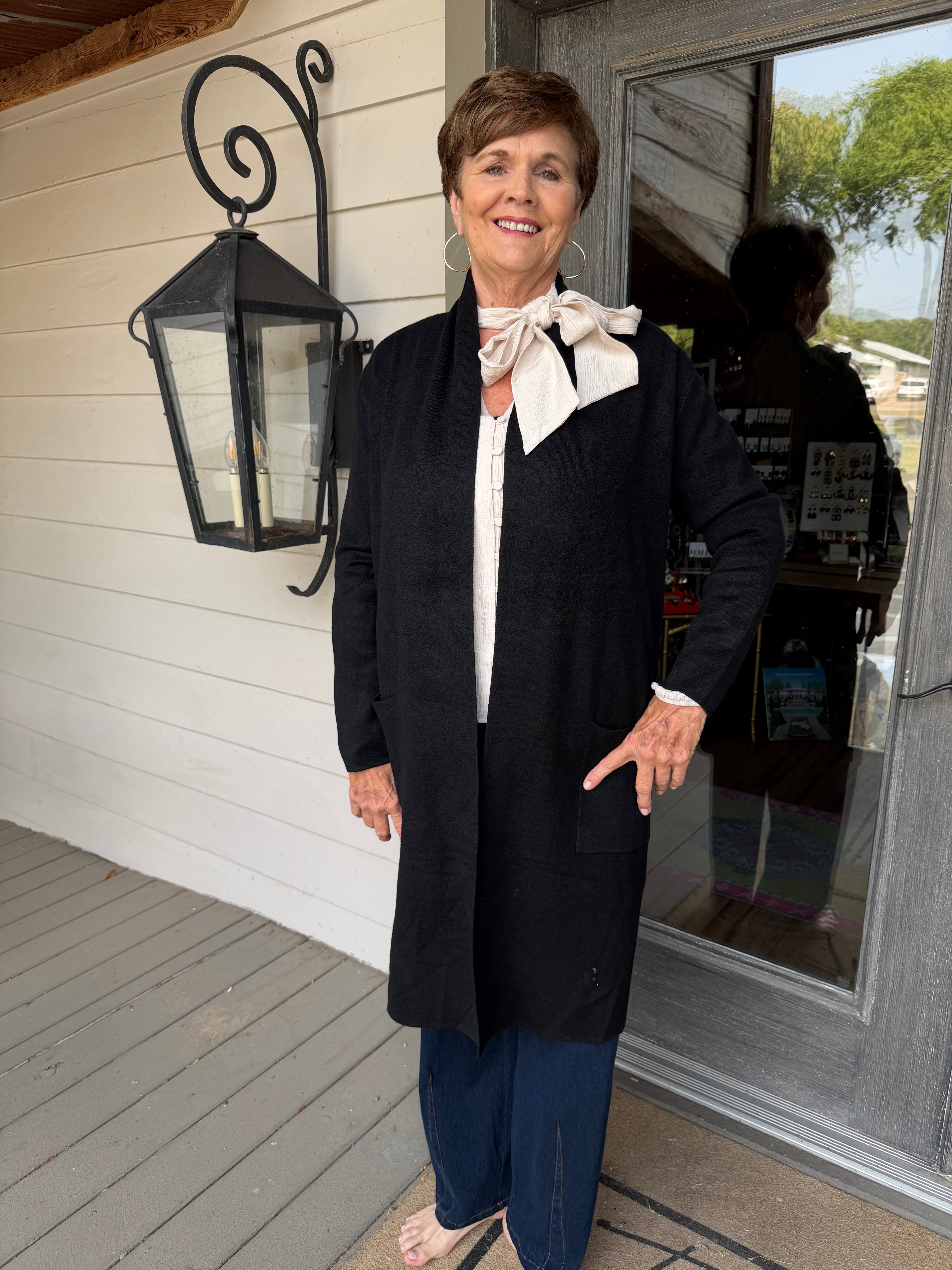 A woman stands on a porch wearing a long black cardigan over a white blouse with a cream bow, navy wide-leg pants, and flat sandals. The background shows a white wood-paneled wall, a black decorative lantern, and a glass door.