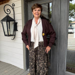 A woman stands on a porch wearing a dark burgundy cardigan, a cream tie-neck blouse, and brown patterned wide-leg pants. She is positioned in front of a gray double door and white siding.