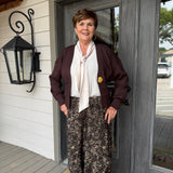 A woman stands on a porch wearing a dark burgundy cardigan, a cream tie-neck blouse, and brown patterned wide-leg pants. She is positioned in front of a gray double door and white siding.