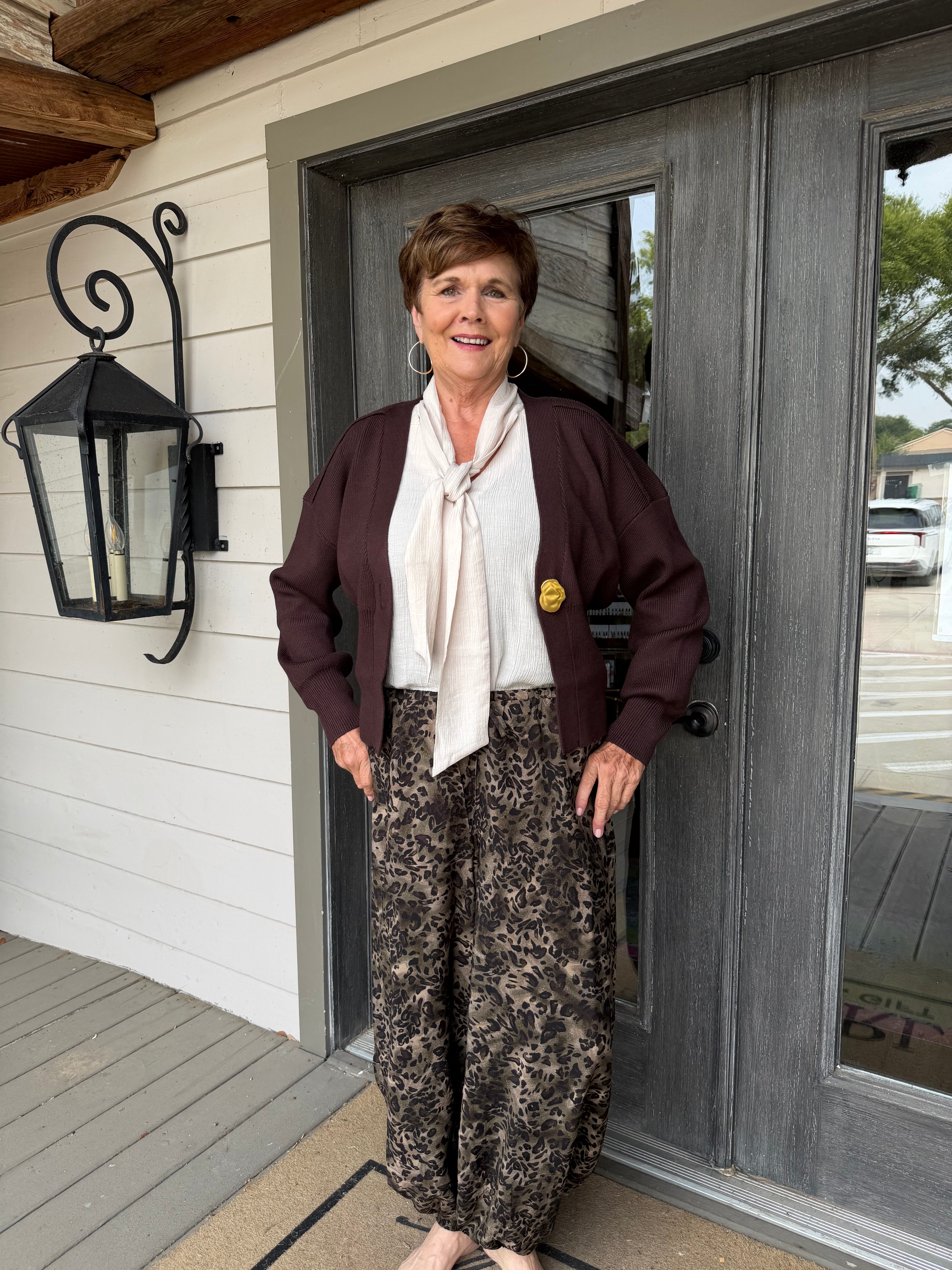 A woman stands on a porch wearing a dark burgundy cardigan, a cream tie-neck blouse, and brown patterned wide-leg pants. She is positioned in front of a gray double door and white siding.