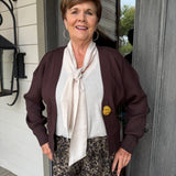 A smiling older woman stands in front of a gray door wearing a dark brown cardigan, a cream tie-neck blouse, and brown patterned pants. She has hoop earrings and a small yellow pin on her cardigan.