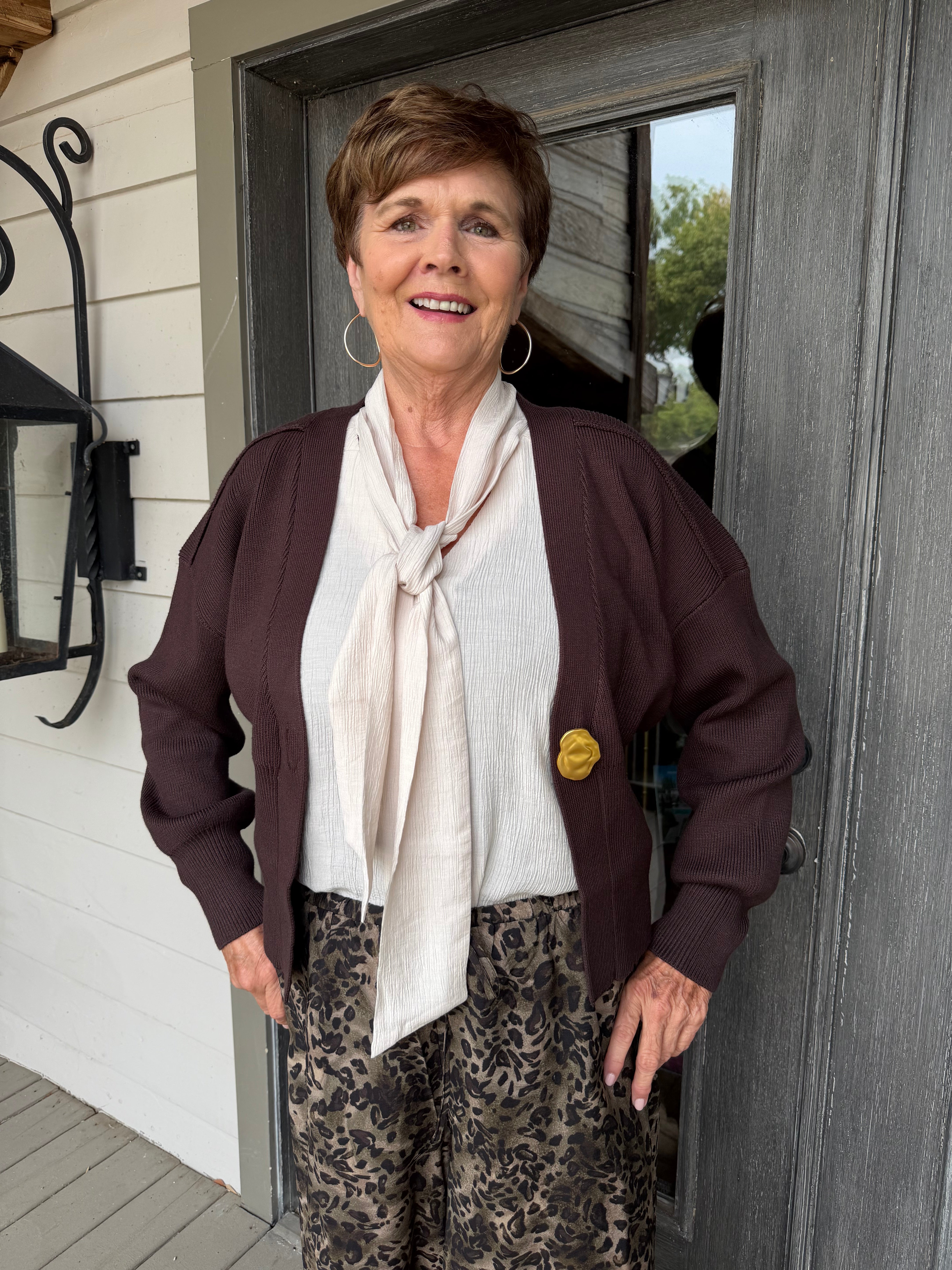 A smiling older woman stands in front of a gray door wearing a dark brown cardigan, a cream tie-neck blouse, and brown patterned pants. She has hoop earrings and a small yellow pin on her cardigan.