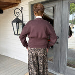 A person viewed from behind wearing a dark plum ribbed knit sweater and wide-leg brown and beige floral print pants, standing on a doormat in front of a gray double door on a porch.