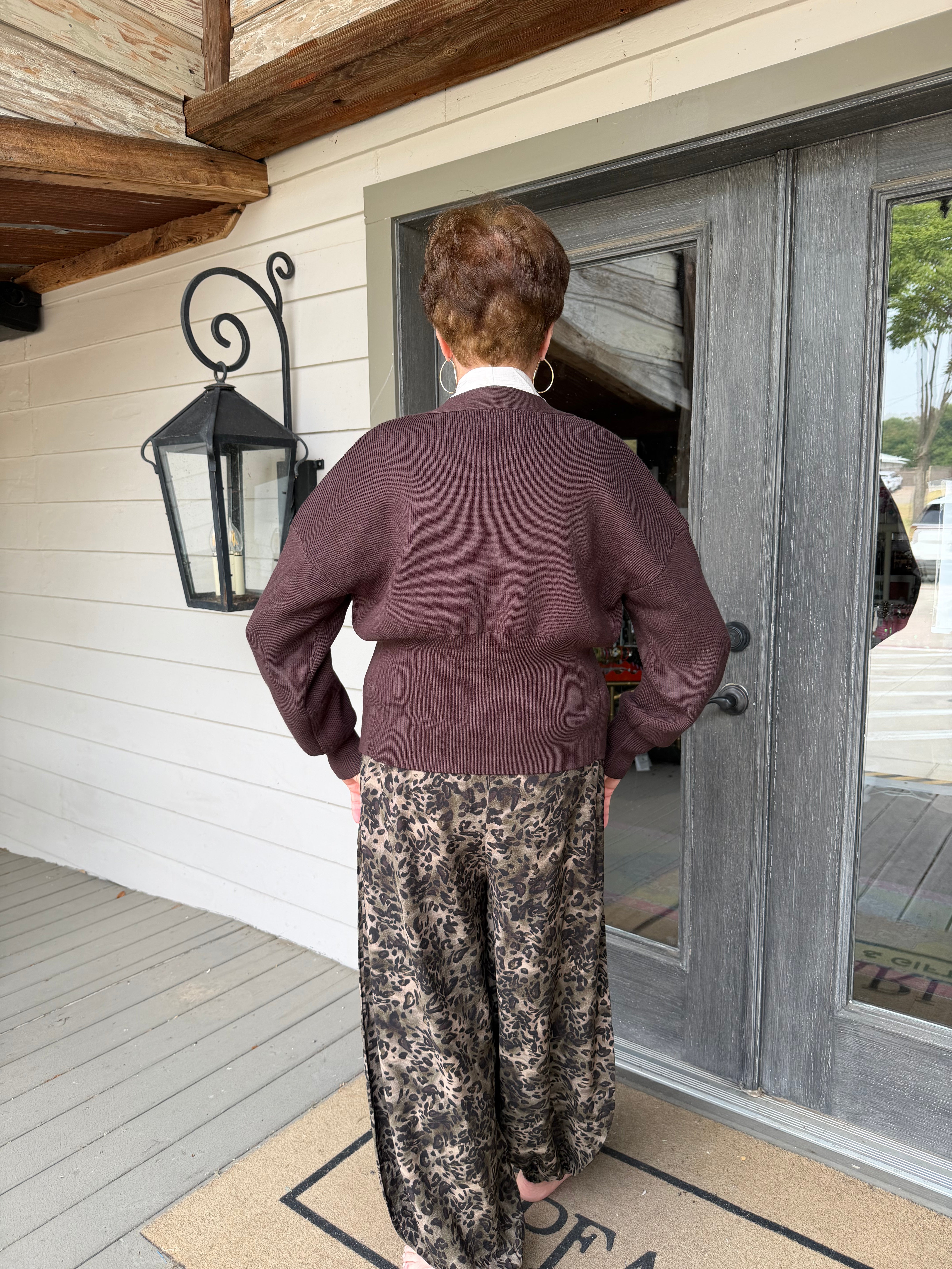 A person viewed from behind wearing a dark plum ribbed knit sweater and wide-leg brown and beige floral print pants, standing on a doormat in front of a gray double door on a porch.