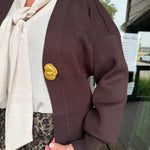 A person wearing a dark brown ribbed knit cardigan with a large textured gold button, layered over a cream tie-neck top and leopard-print pants, standing in front of a storefront window.