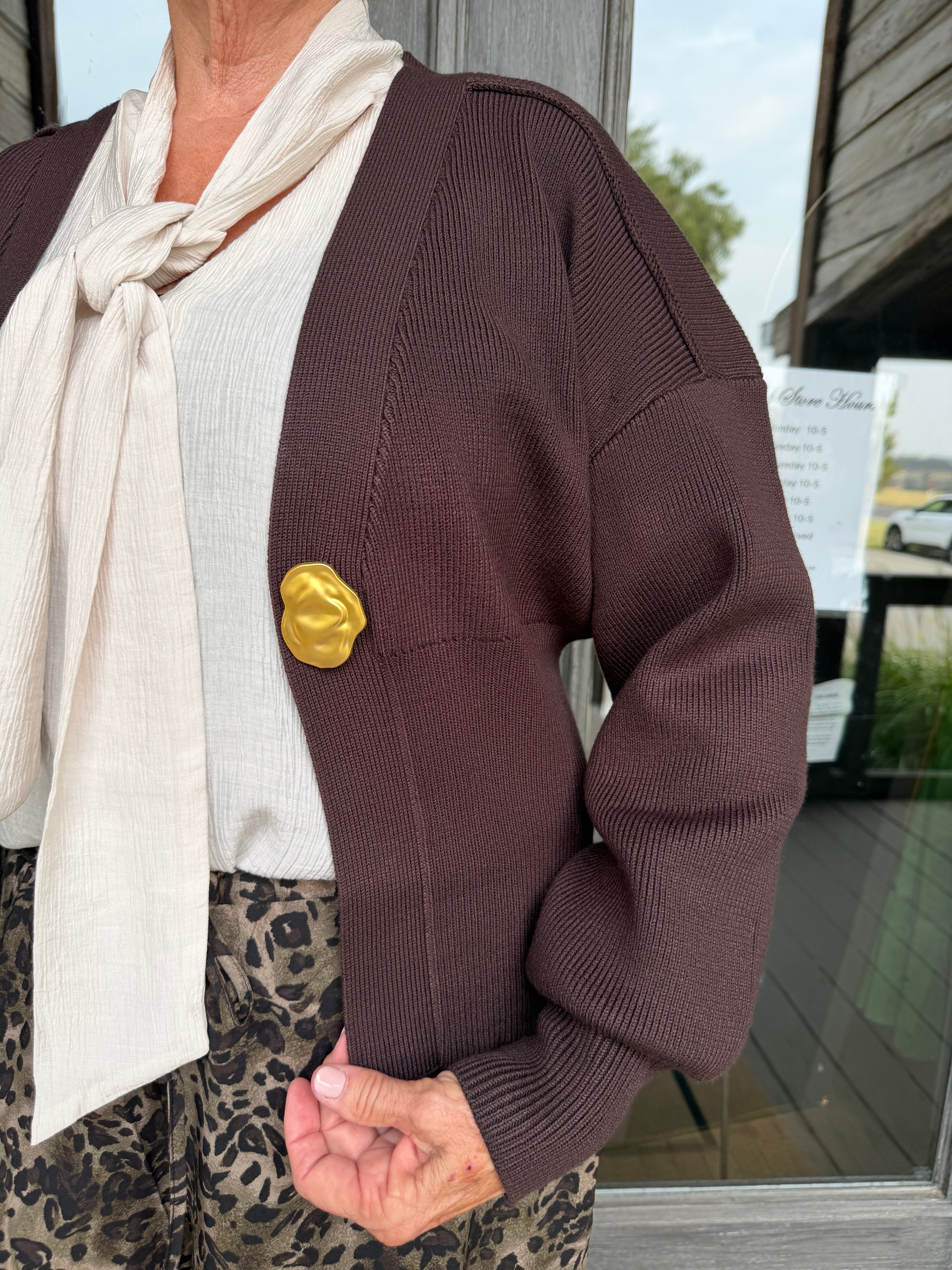 A person wearing a dark brown ribbed knit cardigan with a large textured gold button, layered over a cream tie-neck top and leopard-print pants, standing in front of a storefront window.