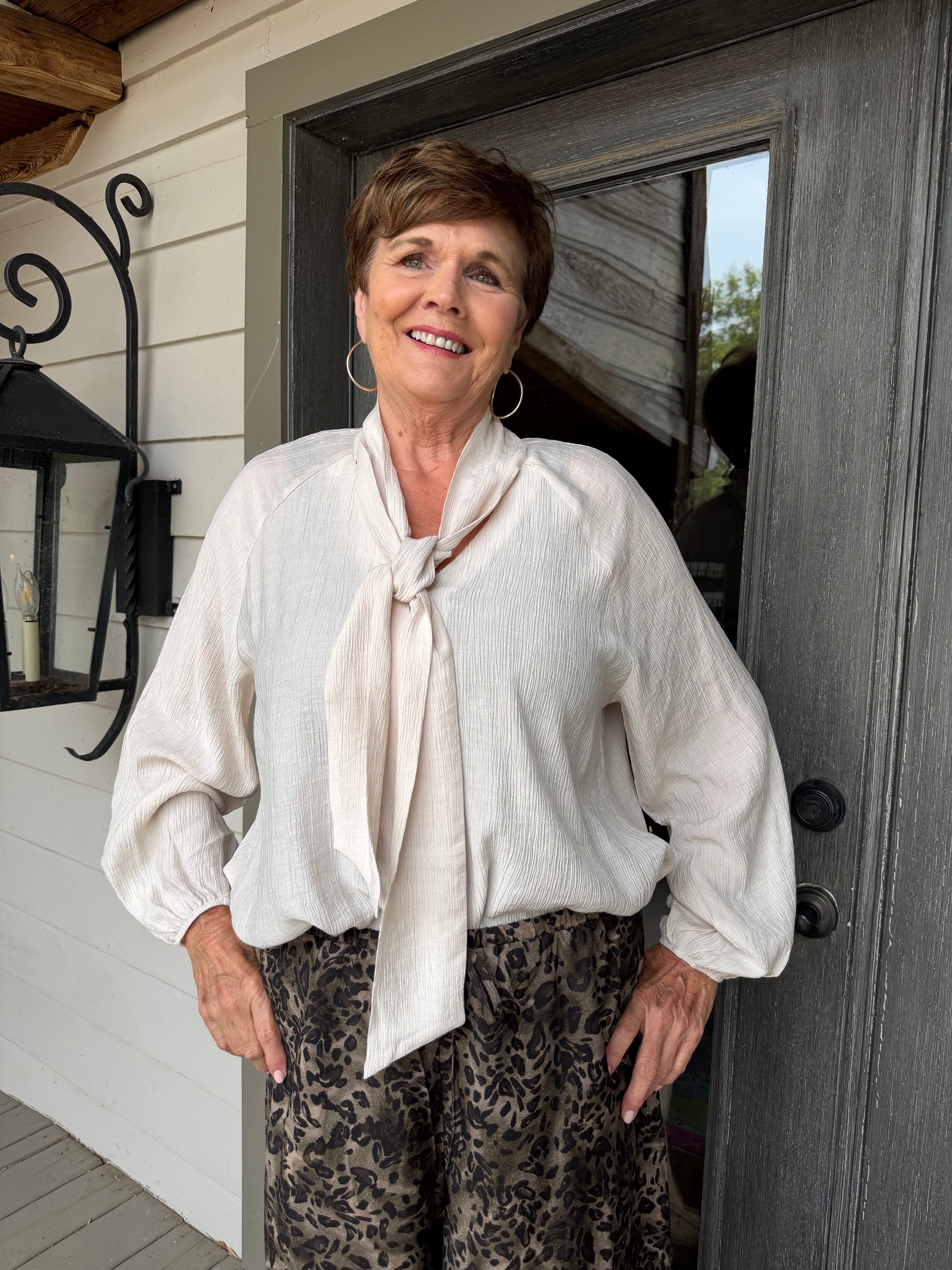 A smiling woman stands on a porch wearing a cream-colored long-sleeve tie-neck blouse and leopard-print pants.