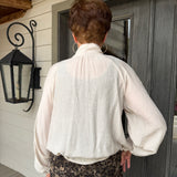A person viewed from behind wearing a cream-colored crinkled blouse with a high gathered neck and long balloon sleeves, paired with brown and black leopard print pants, standing in front of a dark gray door on a porch.