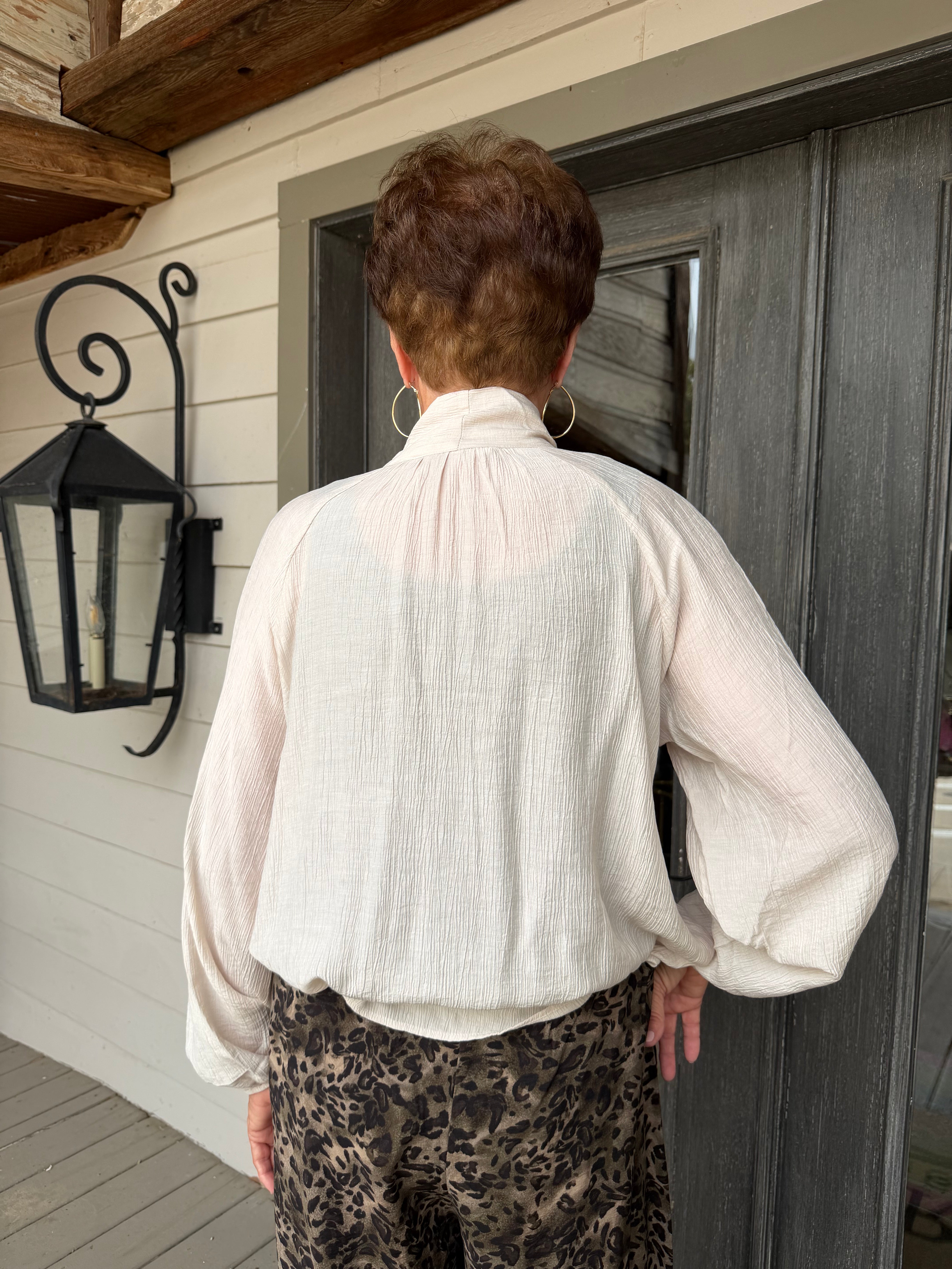 A person viewed from behind wearing a cream-colored crinkled blouse with a high gathered neck and long balloon sleeves, paired with brown and black leopard print pants, standing in front of a dark gray door on a porch.