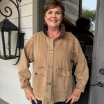 A smiling woman stands on a porch wearing a tan corduroy utility jacket with a brown collar and dark blue pants.