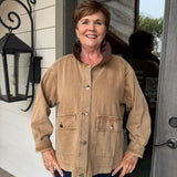 A smiling woman stands on a porch wearing a tan corduroy utility jacket with a brown collar and dark blue pants.