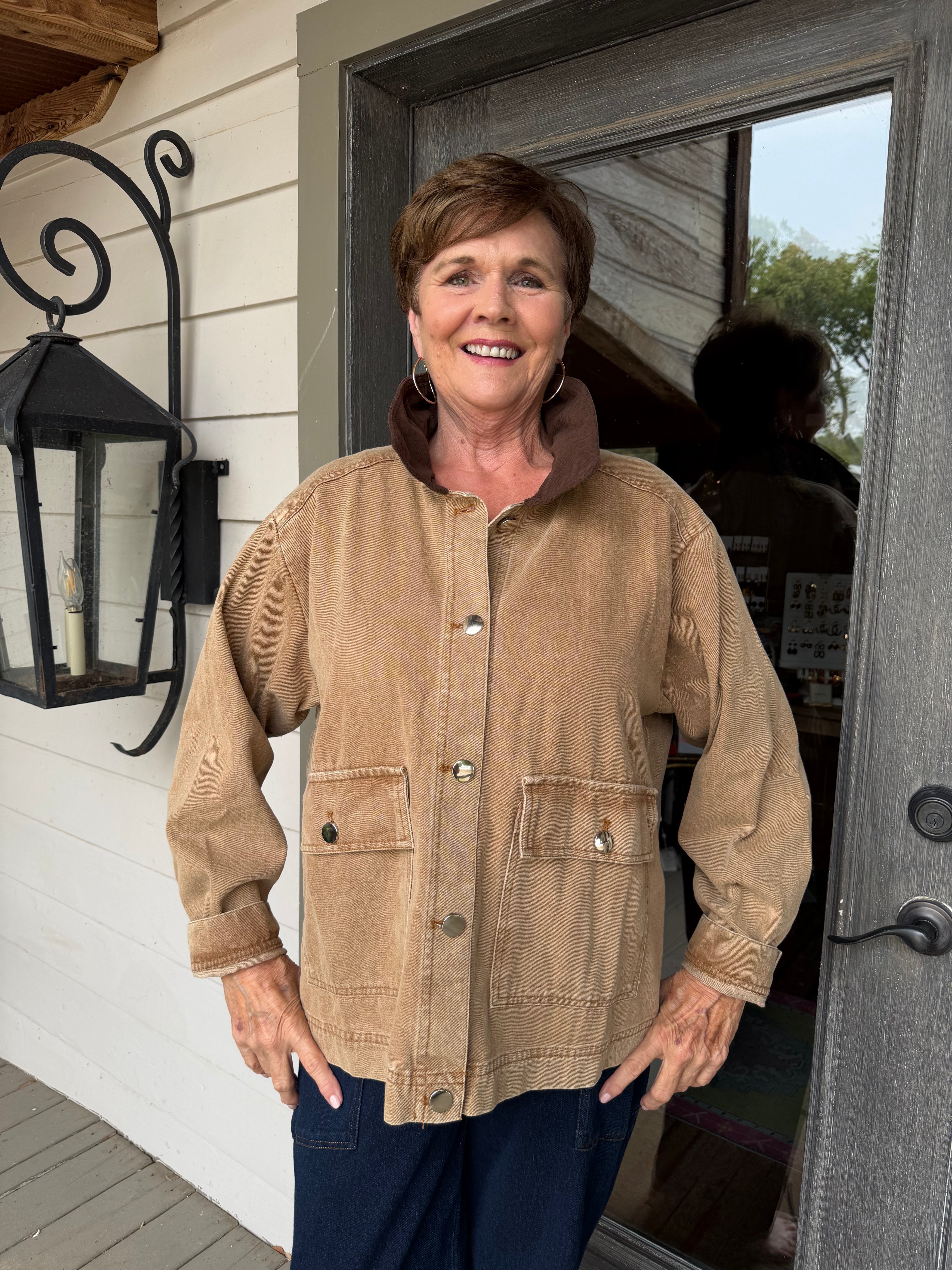 A smiling woman stands on a porch wearing a tan corduroy utility jacket with a brown collar and dark blue pants.