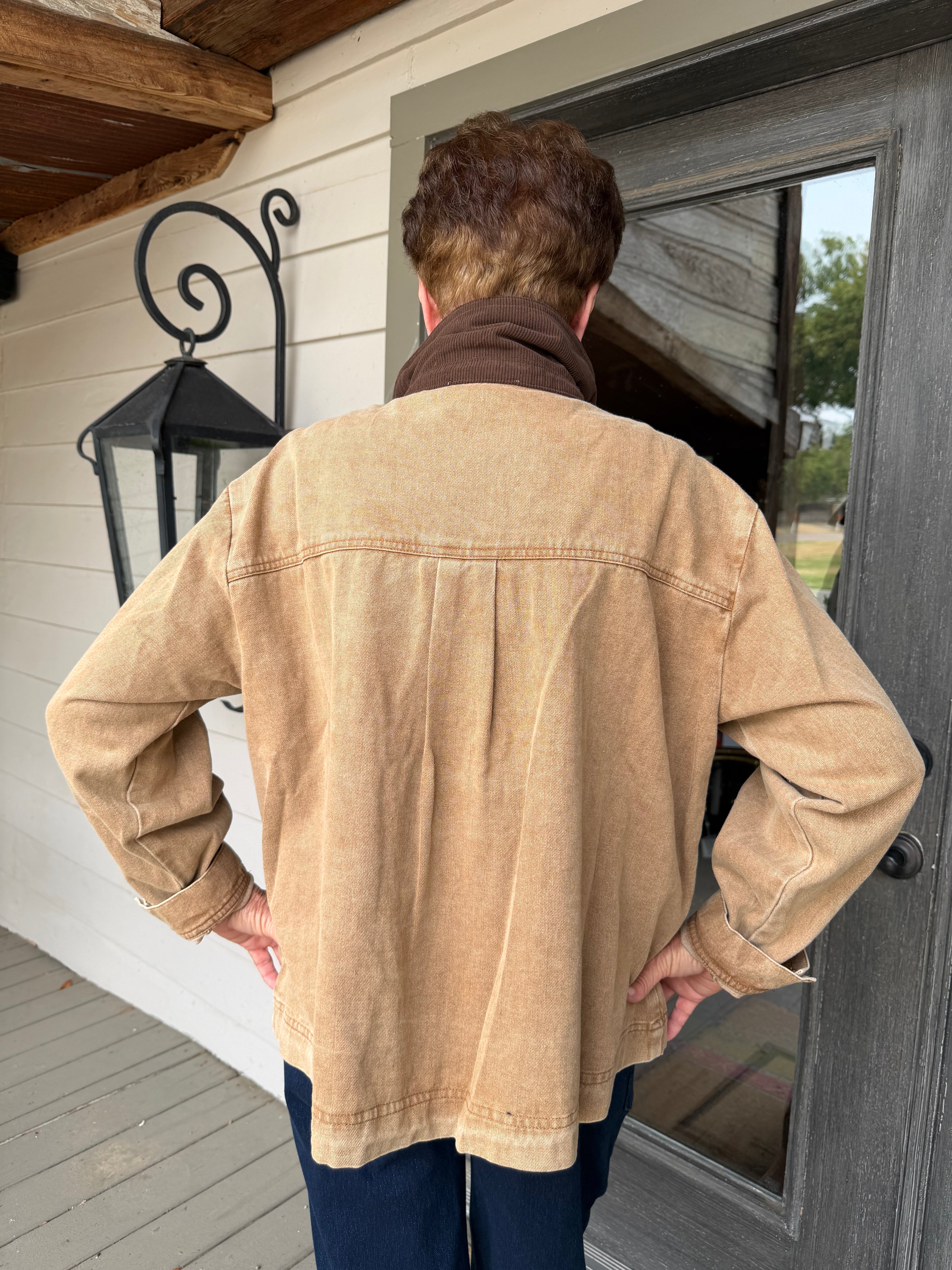 A person viewed from behind wearing a tan canvas utility jacket with a brown collar, standing on a porch in front of a dark gray door.