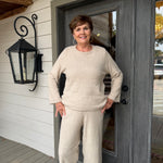 A smiling woman stands on a porch wearing a matching knit two-piece outfit in a light cream or oatmeal color, consisting of a long-sleeve crewneck sweater and wide-leg pants.
