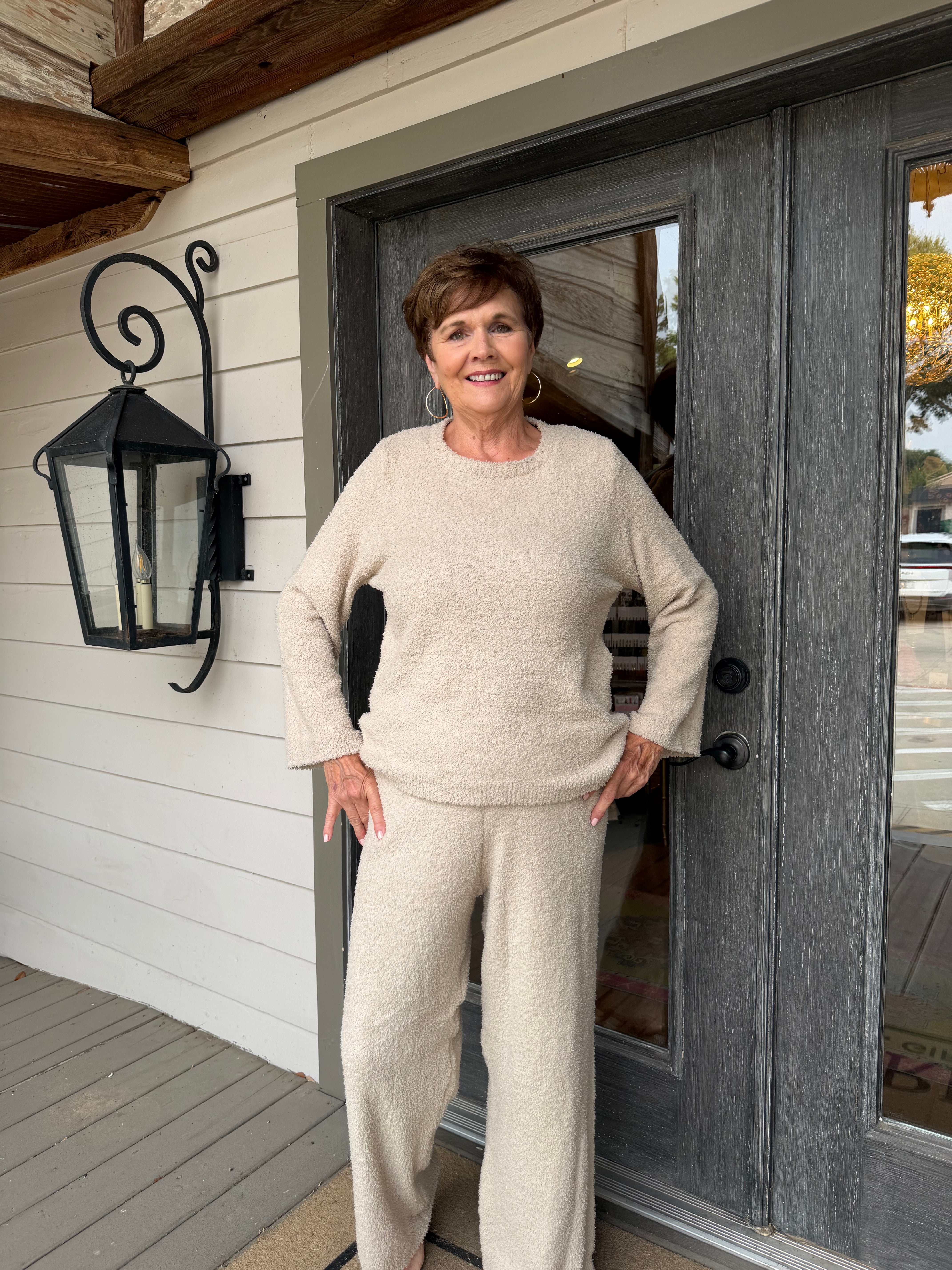 A smiling woman stands on a porch wearing a matching knit two-piece outfit in a light cream or oatmeal color, consisting of a long-sleeve crewneck sweater and wide-leg pants.