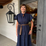 A woman wearing a navy blue midi dress with a brown belt stands on a porch in front of a gray door.
