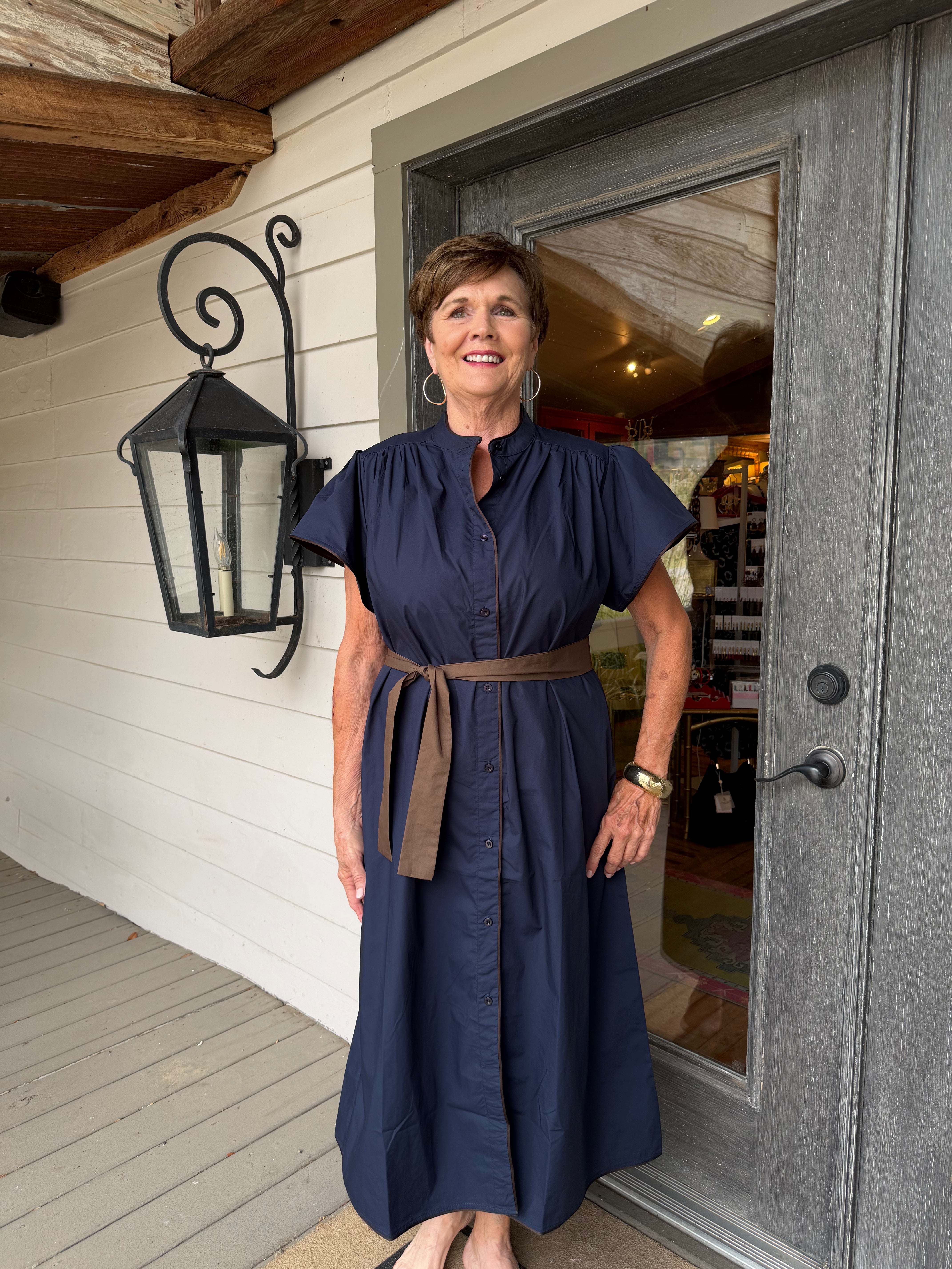A woman wearing a navy blue midi dress with a brown belt stands on a porch in front of a gray door.