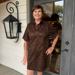 A smiling woman stands on a porch wearing a brown and black leopard-print short-sleeve collared dress. The photo is taken in front of a gray door and white siding, with a black wall lantern visible on the left.