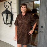 A smiling woman stands on a porch wearing a brown and black leopard-print short-sleeve collared dress. The photo is taken in front of a gray door and white siding, with a black wall lantern visible on the left.