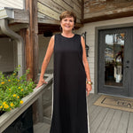 A woman wearing a sleeveless black maxi dress with white side stripes stands on a wooden deck porch in front of a house with wood siding and a glass door.