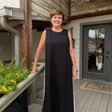 A woman wearing a sleeveless black maxi dress with white side stripes stands on a wooden deck porch in front of a house with wood siding and a glass door.