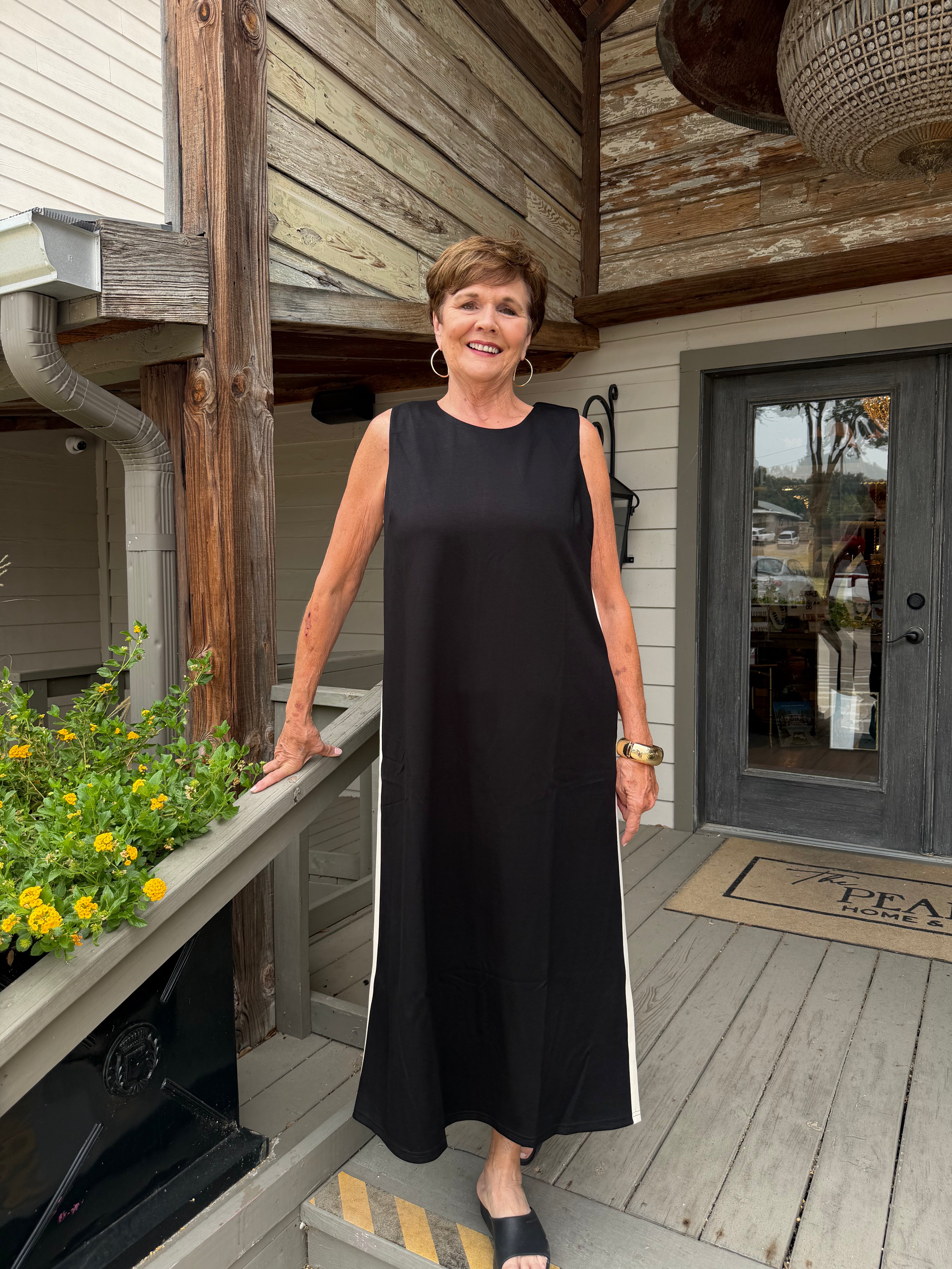 A woman wearing a sleeveless black maxi dress with white side stripes stands on a wooden deck porch in front of a house with wood siding and a glass door.