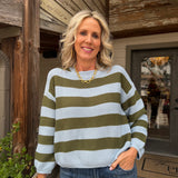 A smiling blonde woman wearing a light blue and olive green horizontally striped knit sweater, blue jeans, and a gold chain necklace, standing on a rustic wooden porch.