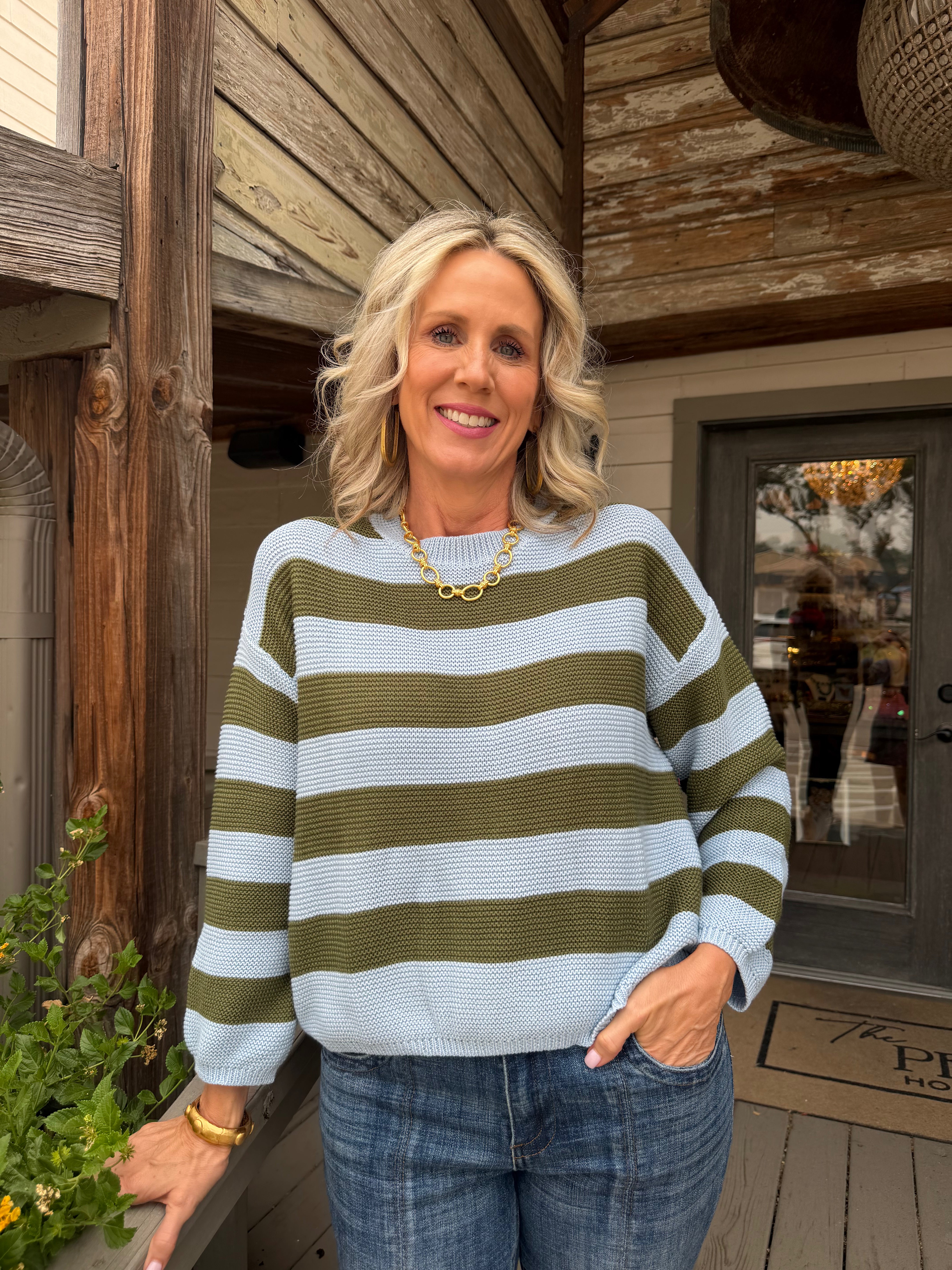 A smiling blonde woman wearing a light blue and olive green horizontally striped knit sweater, blue jeans, and a gold chain necklace, standing on a rustic wooden porch.